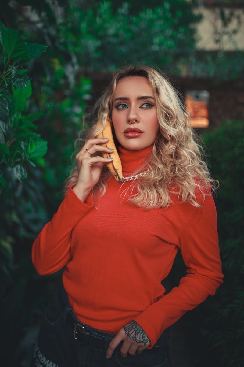 Blonde woman in red poses playfully with banana as phone amidst lush greenery.